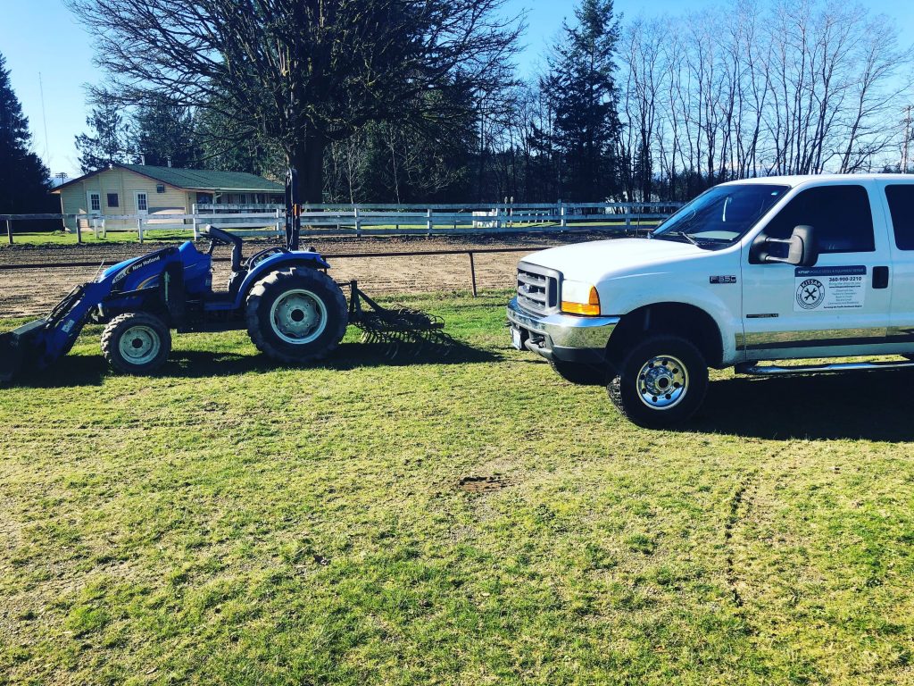 White service pickup truck parked on a grassy property next to a blue tractor with a front loader, with trees and a fence behind